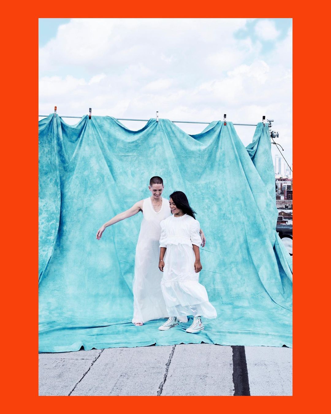 Two women in white dresses pose in front of a blue backdrop outdoors on a sunny day, city buildings visible in the background.