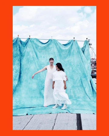 Two women in white dresses pose in front of a blue backdrop outdoors on a sunny day, city buildings visible in the background.