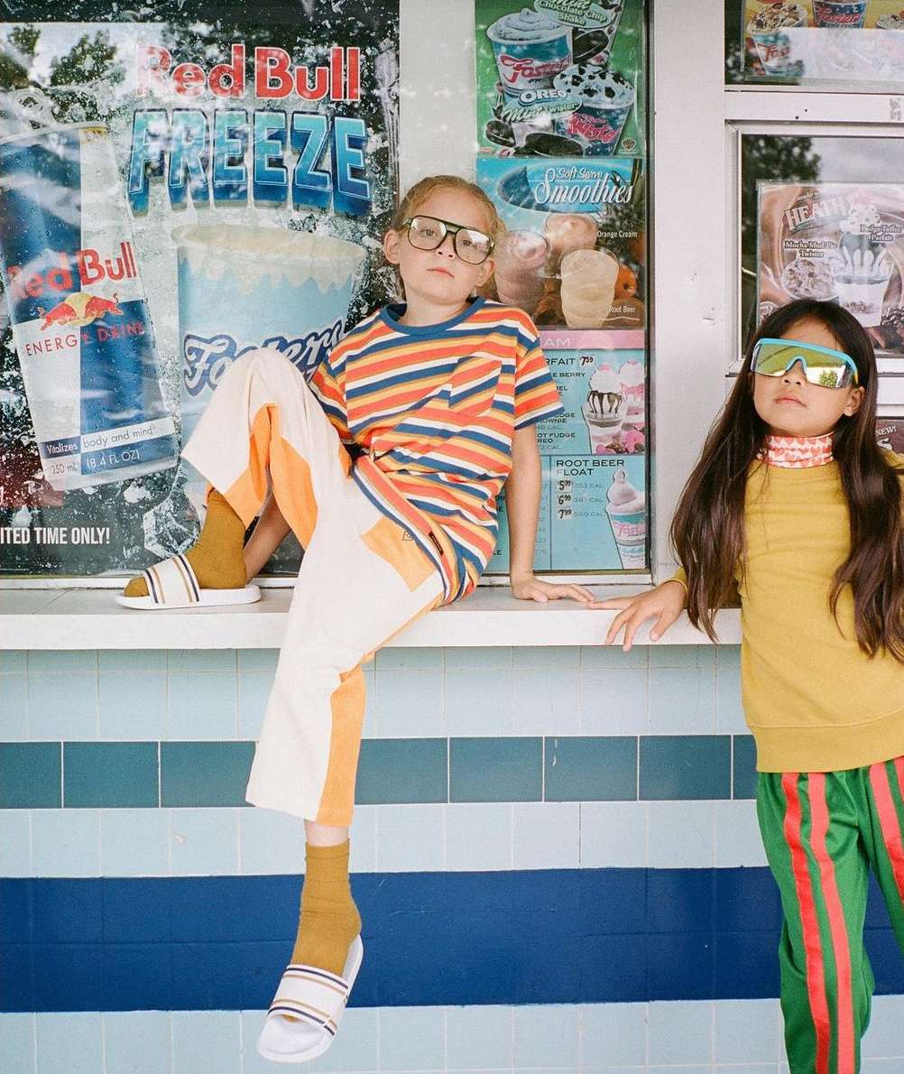 Two children pose in front of a vintage-style advertisement, one seated and wearing a striped shirt and the other standing.