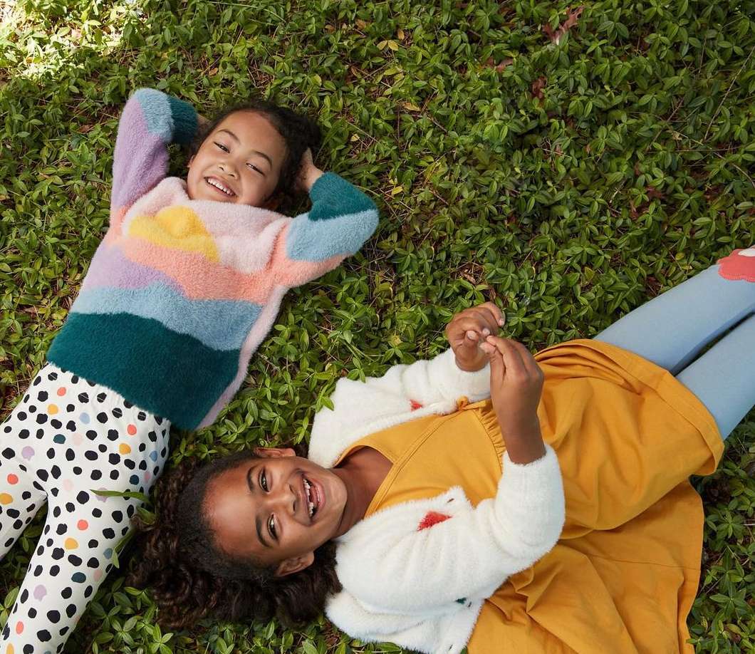 Two smiling girls lie in green grass, one in a colorful striped sweater and polka dot pants, the other in a yellow dress.