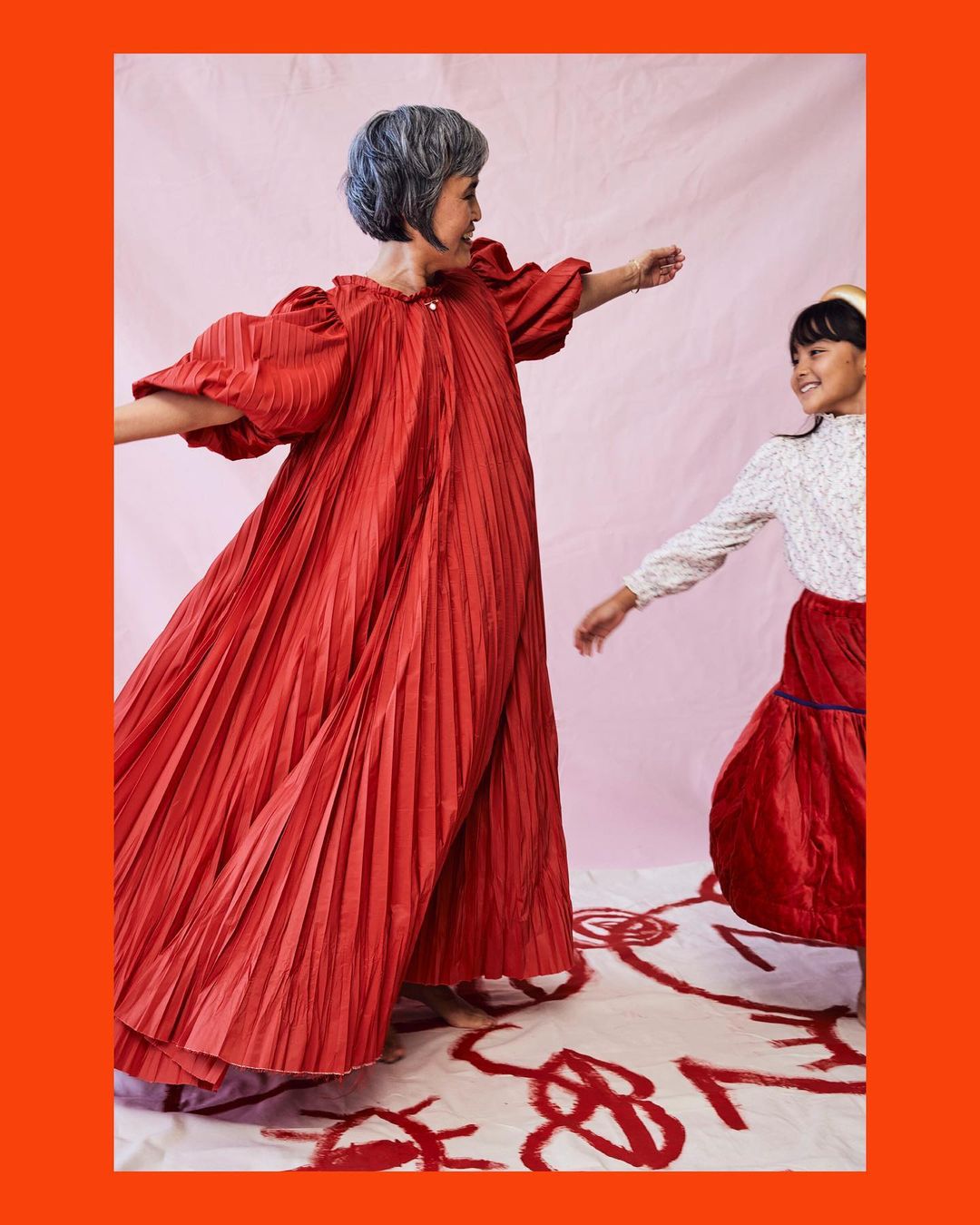 An older adult woman and a child stand on a white surface with red brush strokes, both wearing matching red dresses and looking at each other.