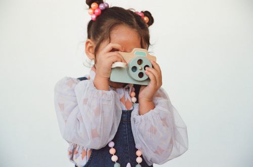 Young child playfully covers face with a toy camera, set against a plain white background. The image has a cute, candid feel.