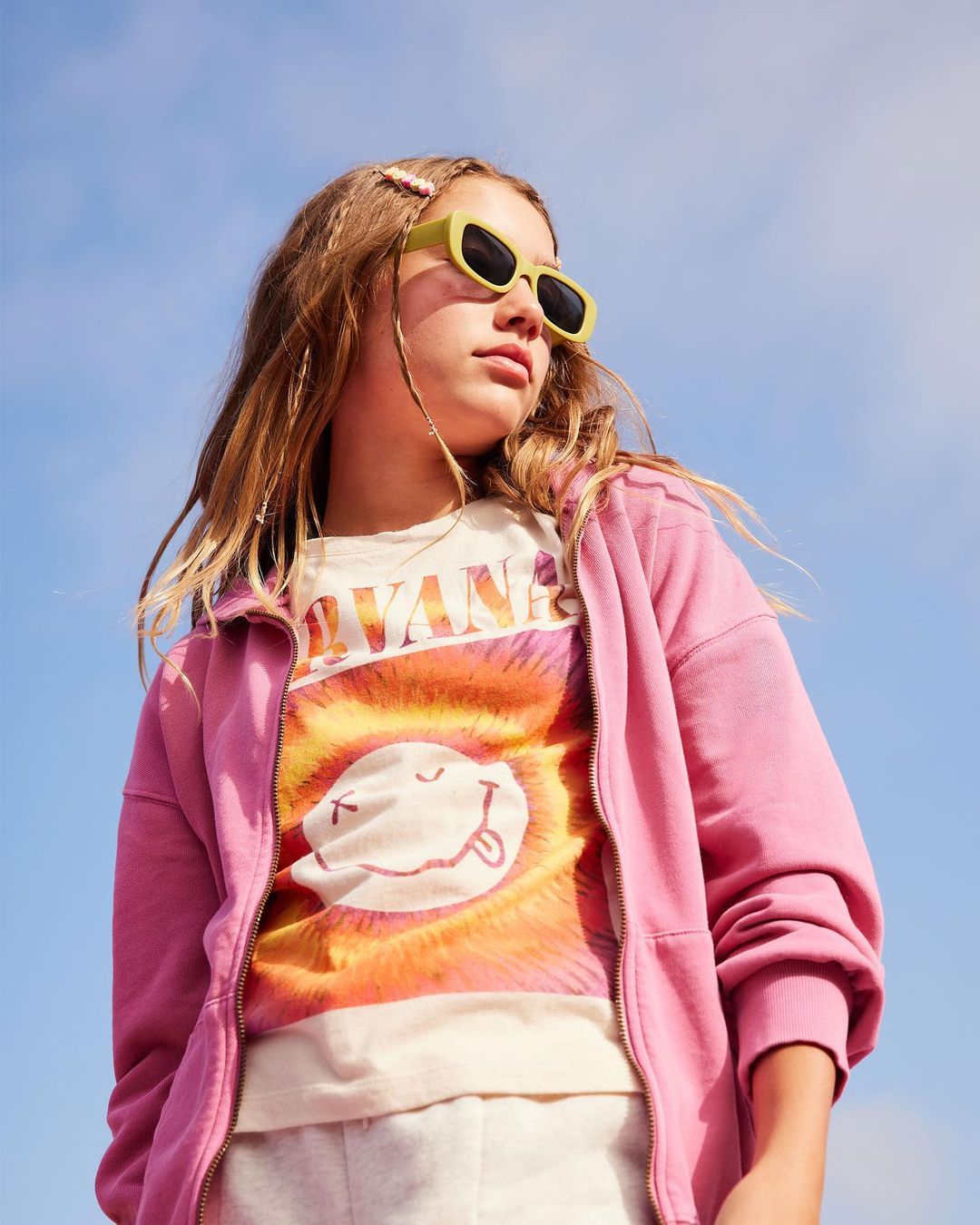 Young girl in pink jacket and lime sunglasses stands against a sunny sky, wearing a vintage graphic tee for a casual lifestyle shot.