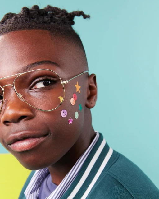 Close-up portrait of a Black child with stickers on their face, wearing gold-rimmed glasses and a varsity jacket against a blue background.