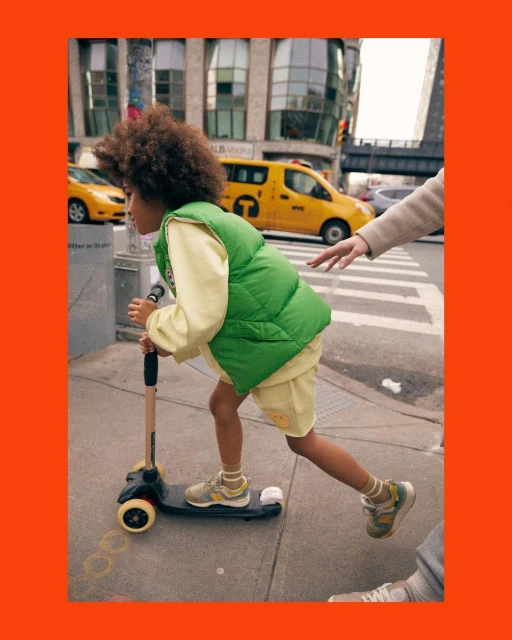 A young child rides a scooter along a sidewalk in a city, wearing a green vest and yellow outfit, with buildings and taxis in the background.