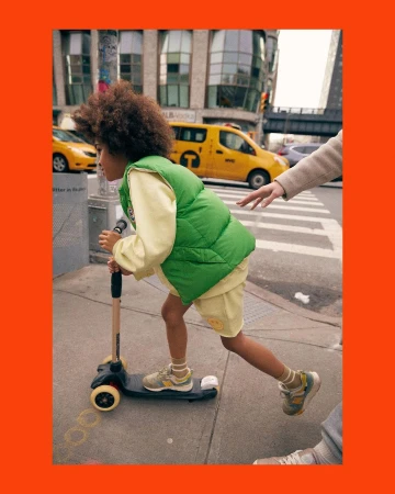 A young child rides a scooter along a sidewalk in a city, wearing a green vest and yellow outfit, with buildings and taxis in the background.