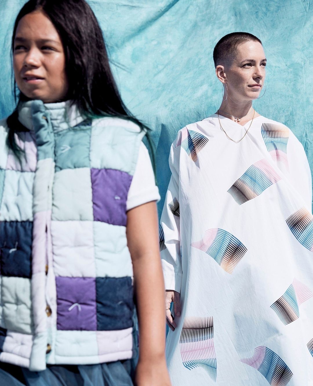 Two women pose indoors with a blue backdrop; one wears a patchwork vest, the other a patterned dress, in soft, diffused lighting.