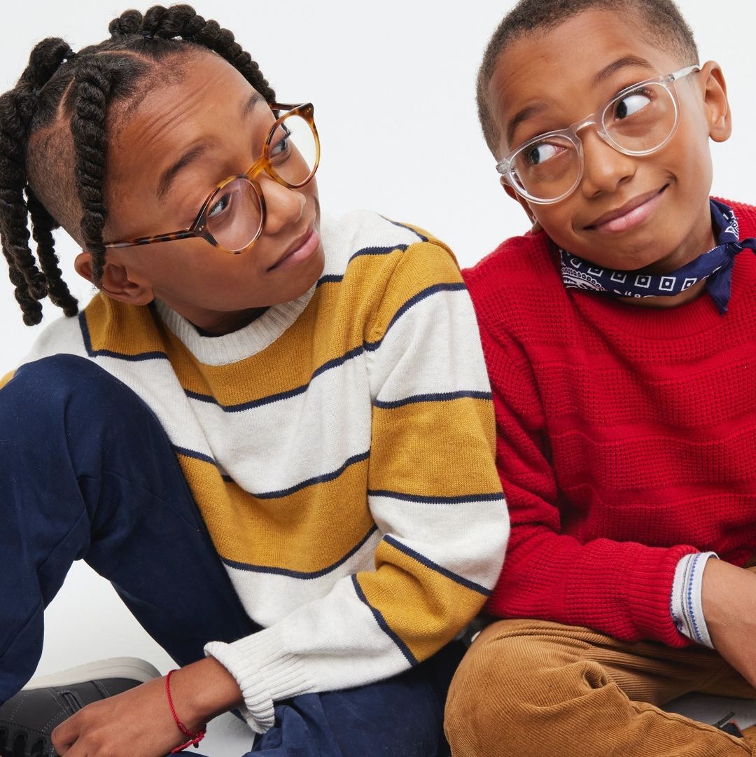 Two young boys in eyeglasses, seated indoors wearing casual sweaters; one in yellow stripes and the other in red, against a bright background.