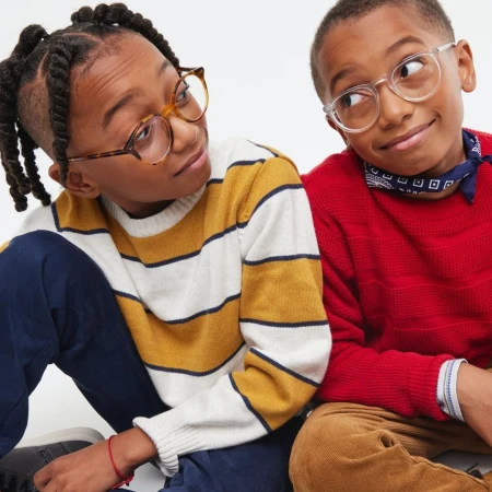 Two young boys in eyeglasses, seated indoors wearing casual sweaters; one in yellow stripes and the other in red, against a bright background.