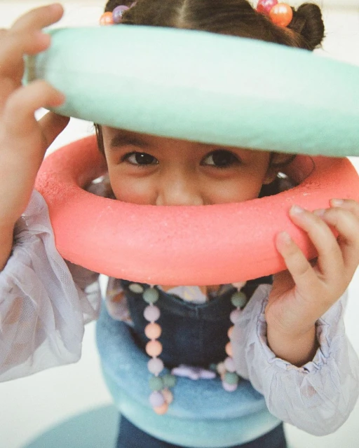 Child with colorful beaded hair and necklace looks through pink and green toy rings in an eye-level, medium indoor shot.
