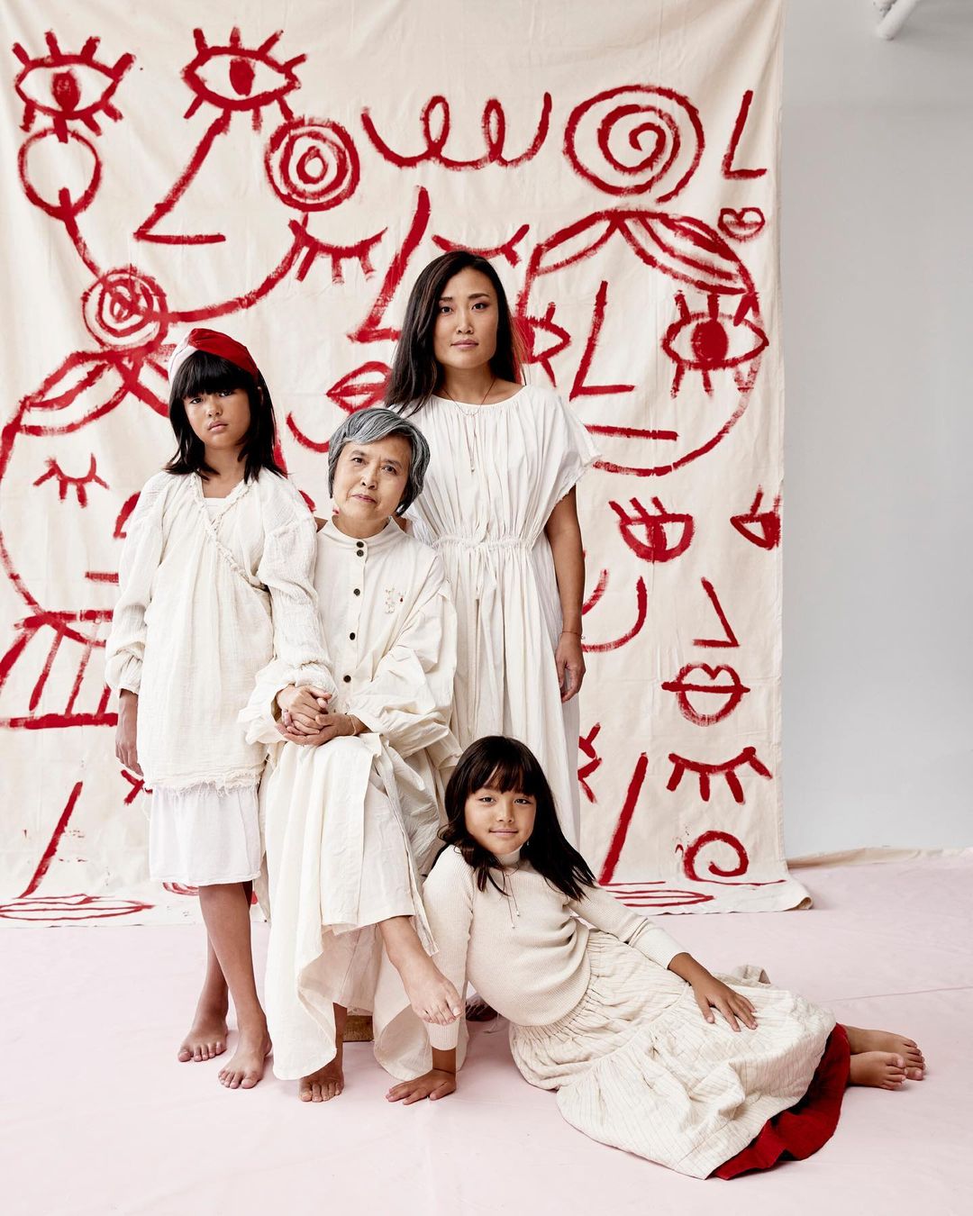 Four Asian women and girls in white dresses pose indoors against a backdrop with a red abstract faces design.