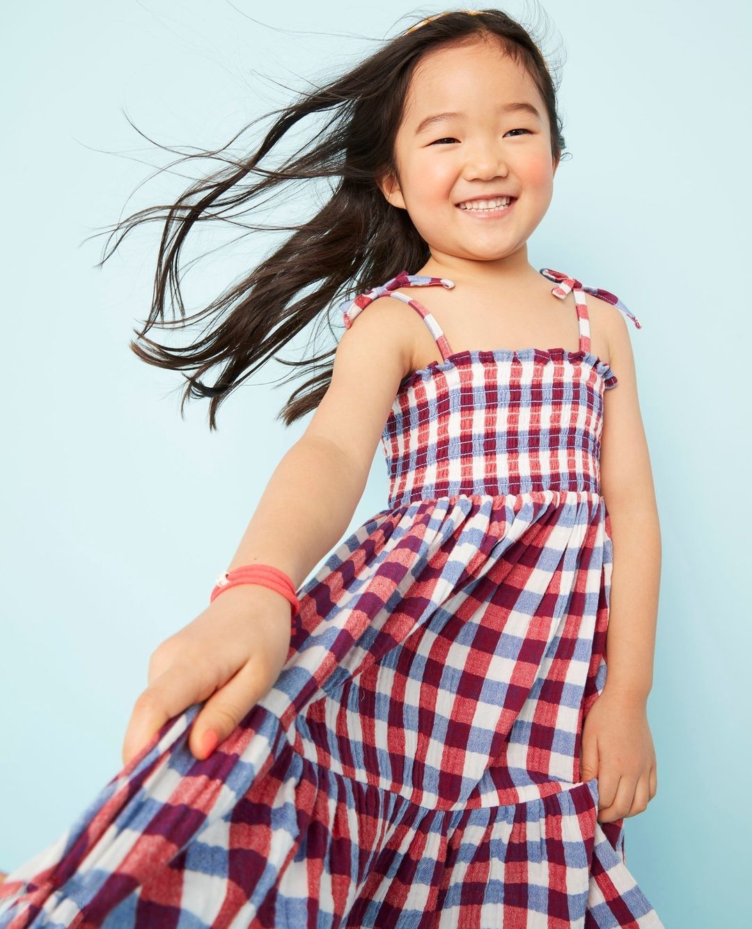 Smiling Asian child twirls in a red and white checkered dress against a blue backdrop, capturing a carefree and joyous moment.