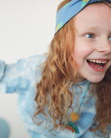 A happy red-haired child with a blue headband and a graphic sweatshirt smiles, with a rainbow necklace, in a brightly lit studio.