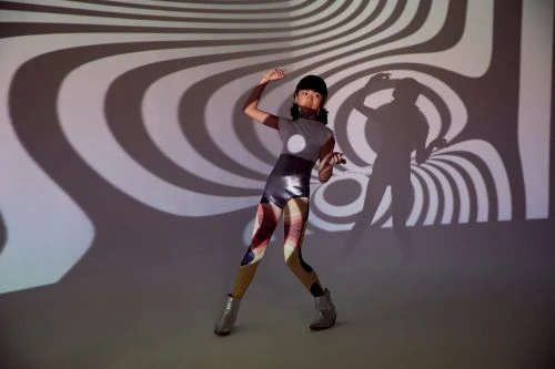 Young female fashion model poses in a studio with swirling black and white pattern projections, wearing a futuristic bodysuit and ankle boots.