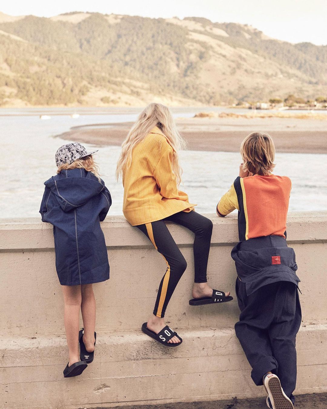 Three children are sitting on a concrete wall overlooking a bay with rolling hills in the distance on a bright, sunny day.
