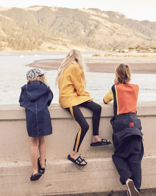 Three children are sitting on a concrete wall overlooking a bay with rolling hills in the distance on a bright, sunny day.
