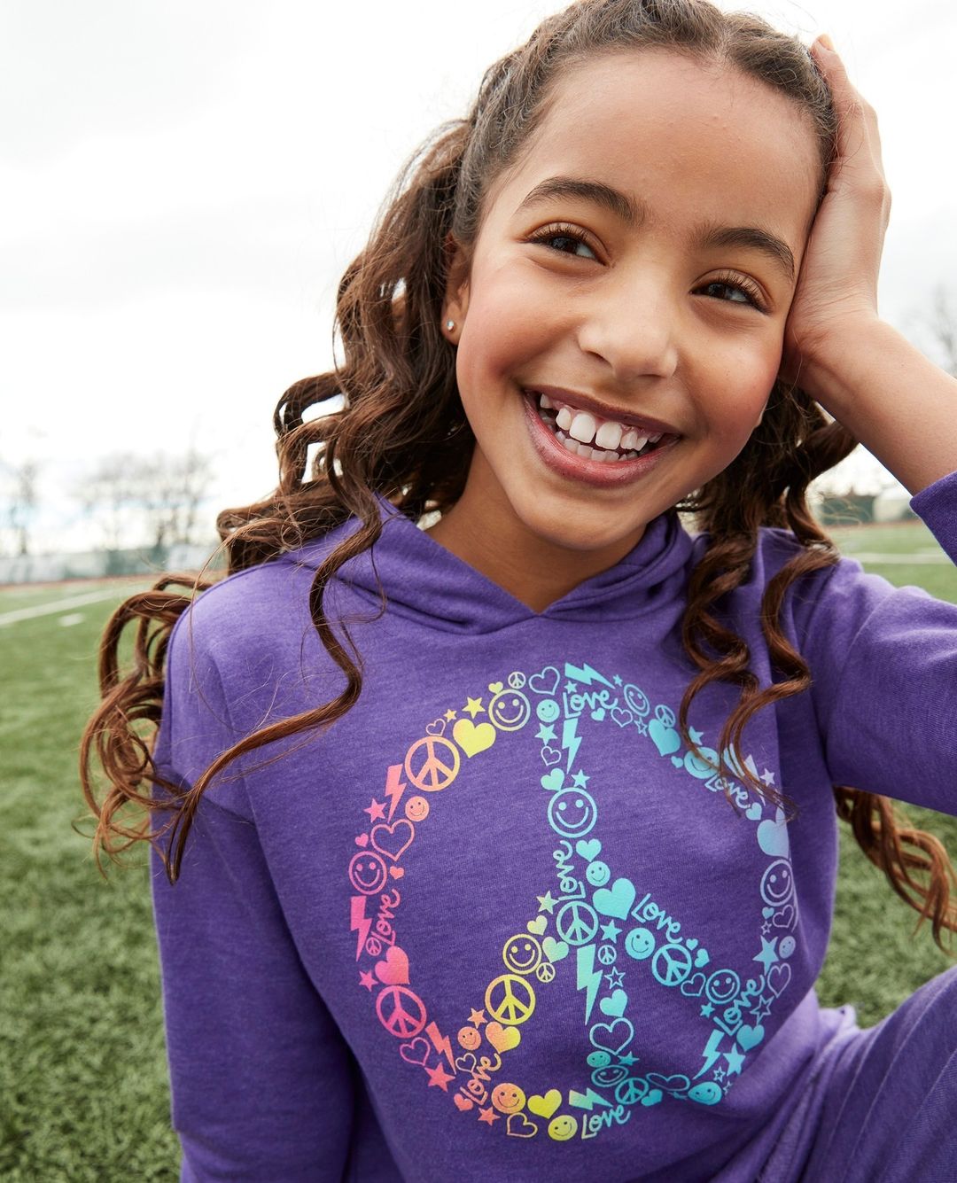 Smiling tween girl in purple peace-sign hoodie, outdoors on a grassy field. Head-and-shoulders shot, eye-level view.