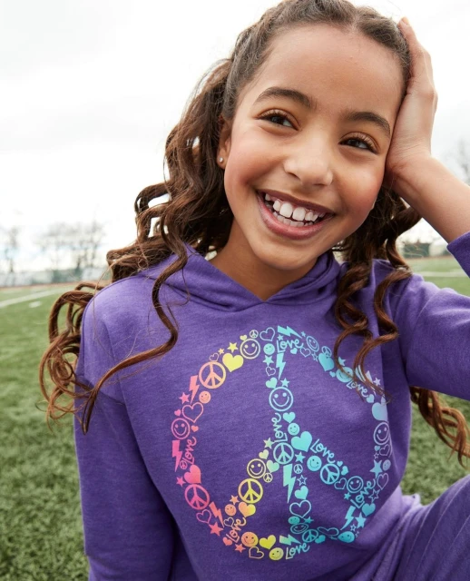 Smiling tween girl in purple peace-sign hoodie, outdoors on a grassy field. Head-and-shoulders shot, eye-level view.