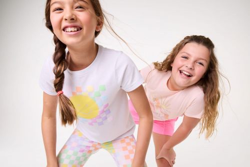 Two smiling girls pose indoors. One girl with braided hair wears a white shirt and colorful leggings; the other wears pink. 