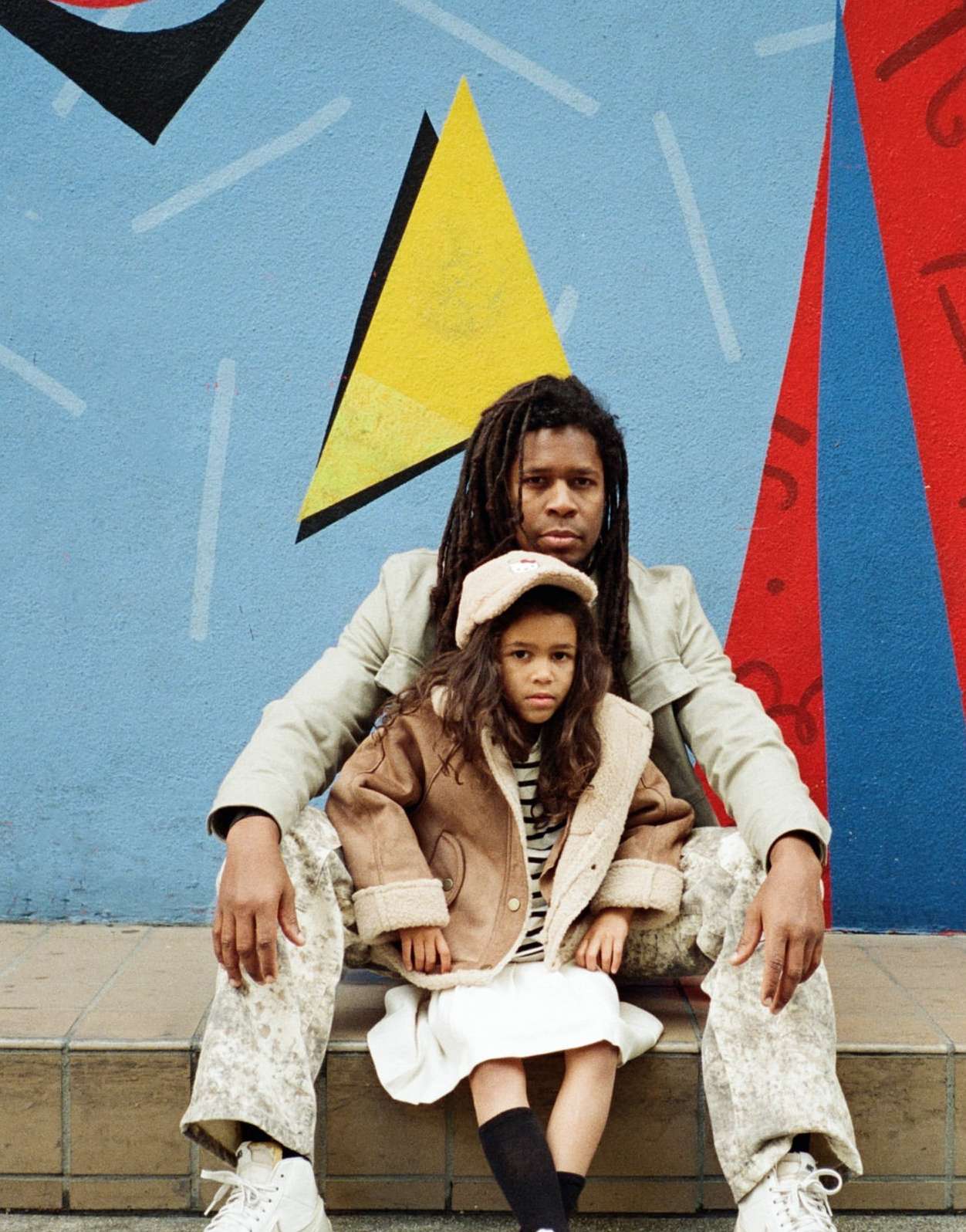 A man with dreadlocks sits on stone steps with a little girl wearing a tan sherpa jacket, in front of a colorful geometric painted wall.