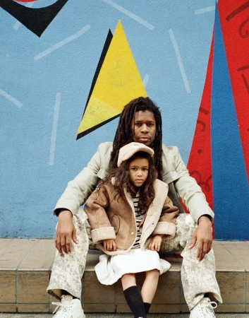 A man with dreadlocks sits on stone steps with a little girl wearing a tan sherpa jacket, in front of a colorful geometric painted wall.