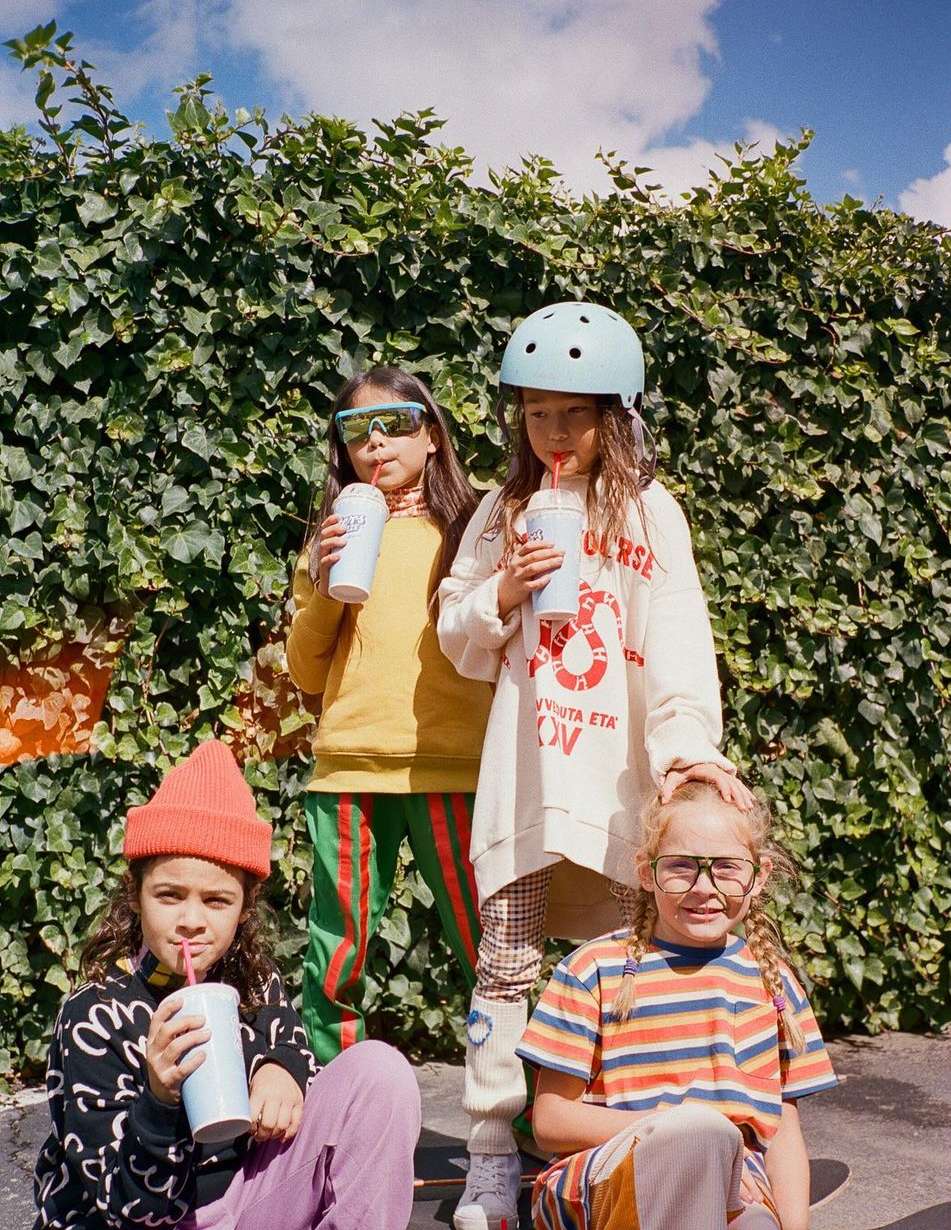 Four young children pose outdoors with drinks and colorful, patterned clothing in front of a green hedge under a partly cloudy sky.