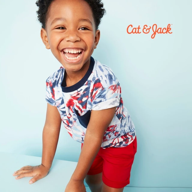A smiling young Black child in a colorful tie-dye style shirt and red shorts stands near a blue surface in a studio shot.