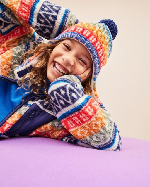A smiling child wearing a colorful patterned fleece jacket and hat reclines on a purple surface in a close-up studio shot.