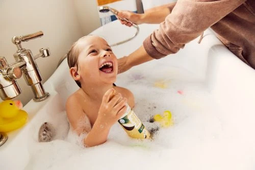 A smiling child with bubbly bathwater, holding a bottle, is being rinsed by an adult in a well-lit bathroom with rubber ducks.