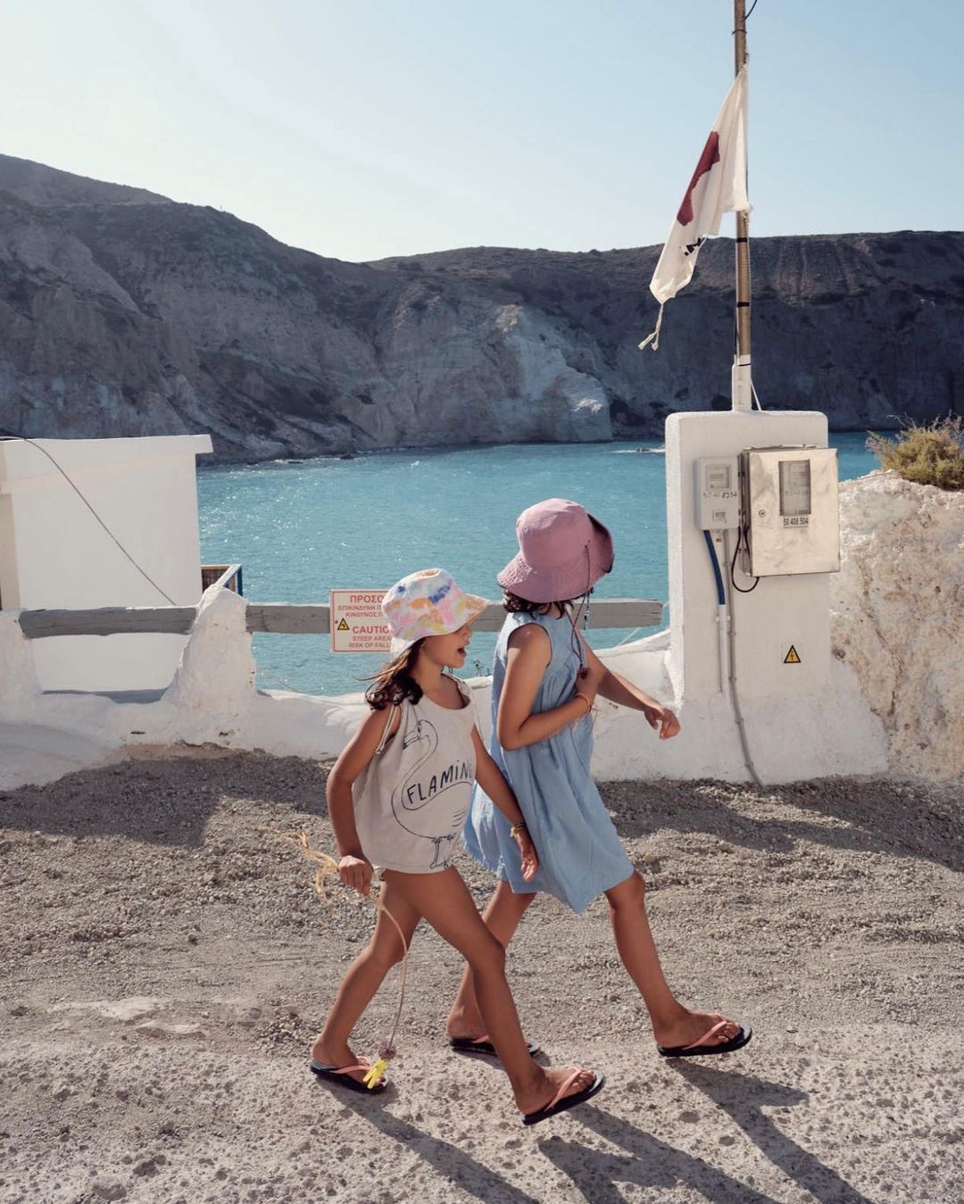 Two young girls in dresses and hats walk along a coastal path, with a view of the blue sea and rocky cliffs in the background.