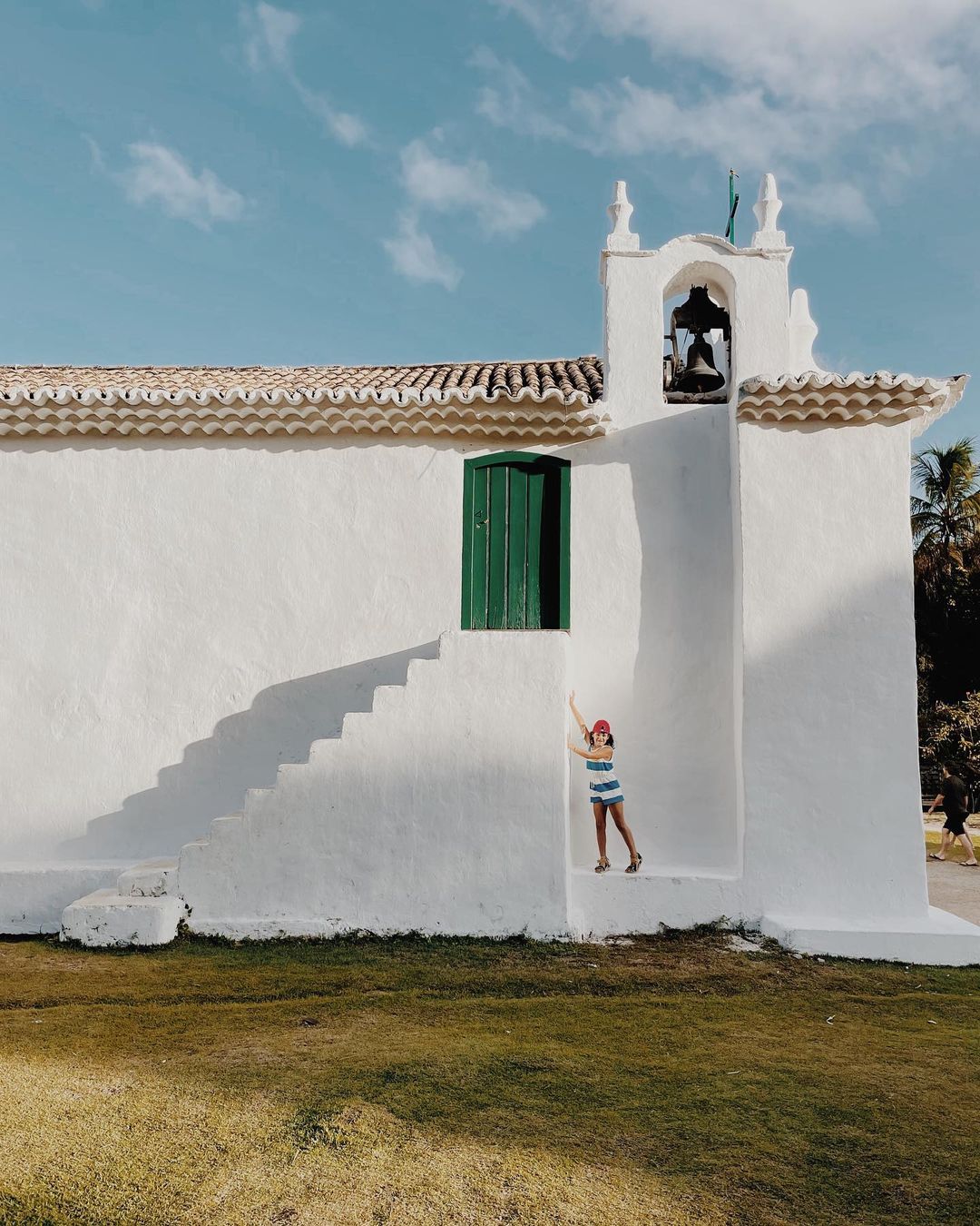 Young girl poses near white chapel stairs under blue sky. Child in striped shirt stands at chapel exterior with lush green lawn.
