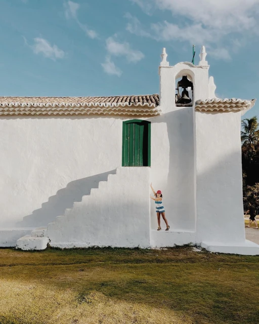 Young girl poses near white chapel stairs under blue sky. Child in striped shirt stands at chapel exterior with lush green lawn.