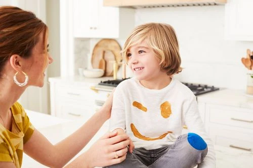 Mother and child in a bright, modern kitchen. The child is smiling and sitting on the kitchen counter. The mother is holding the child.