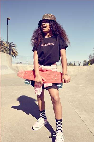 Young girl with curly hair wearing a camo bucket hat, black tee, and Vans sneakers posing outdoors with an orange skateboard.