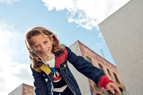 Child with long blonde hair wearing a denim jacket outdoors against a clear blue sky and buildings.