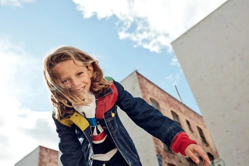 Child with long blonde hair wearing a denim jacket outdoors against a clear blue sky and buildings.