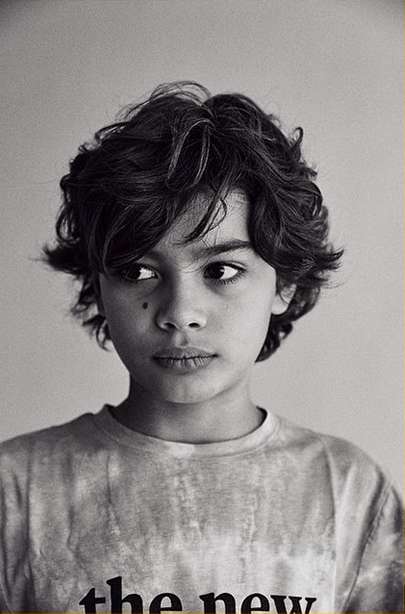 Black and white portrait of a boy with dark curly hair wearing a t-shirt, photographed indoors with natural lighting.