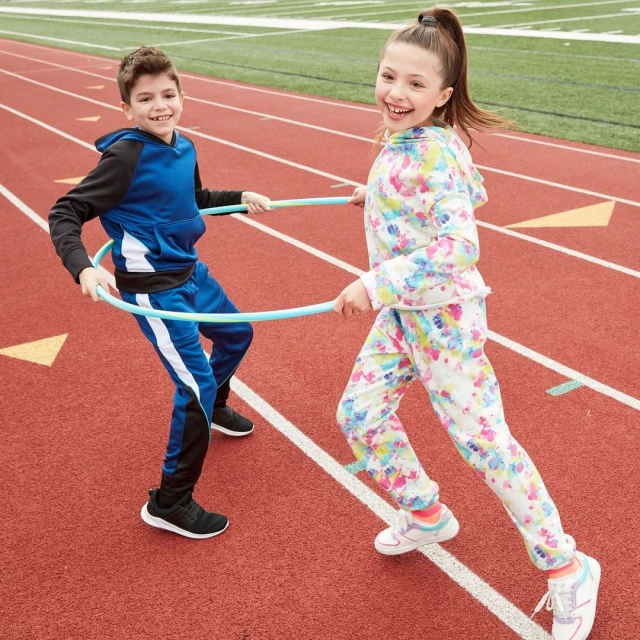 Two smiling children, a boy and a girl, play with hula hoops on a red running track on a sunny day, showcasing active lifestyle and childhood fun.