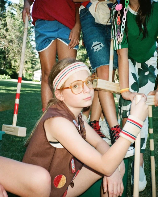 Portrait of a child in vintage brown vest and large glasses with friends holding croquet mallets on a grassy lawn on a sunny day.