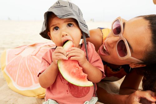 A baby in a sun hat eating a watermelon slice on a beach with a smiling adult woman in sunglasses in the background.