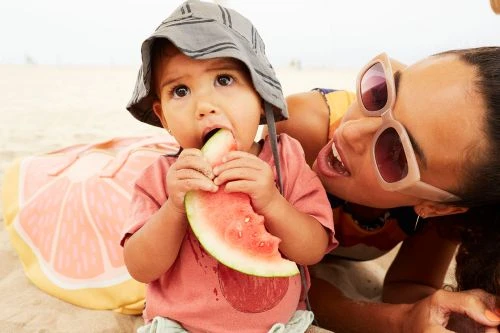 A baby in a sun hat eating a watermelon slice on a beach with a smiling adult woman in sunglasses in the background.