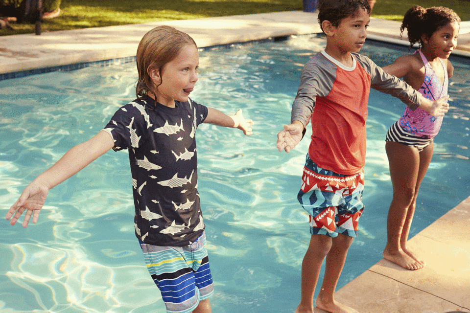 Three children stand on the edge of an outdoor pool, ready to jump in. One boy wears a shark shirt, and the other wears a red shirt.