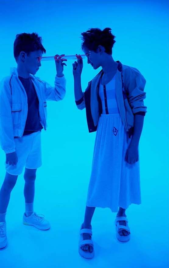Two children in a blue-lit studio hold a glass tube, both dressed in white outfits, face each other in a vibrant and minimalist scene.