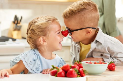 A smiling girl and boy with a strawberry on their noses in a bright, indoor kitchen setting with bowls of fresh strawberries.