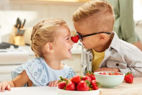 Two young children, a boy and a girl, playfully touch noses with a strawberry between them, laughing in a bright kitchen setting.