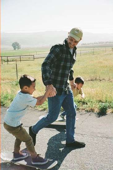 A father holds the hand of his son on a skateboard while another child walks beside them on a paved road with a grassy, hilly landscape in the background.