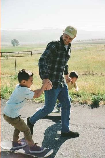 A father holds the hand of his son on a skateboard while another child walks beside them on a paved road with a grassy, hilly landscape in the background.