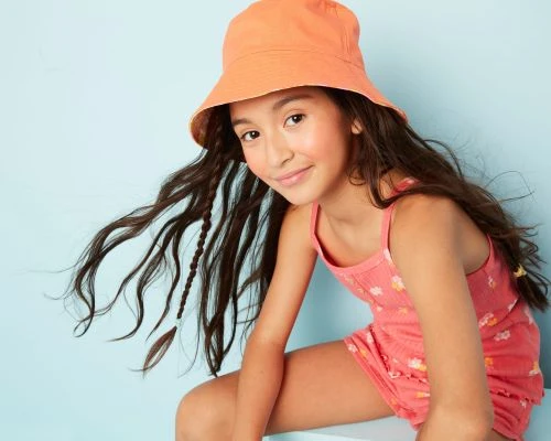 Portrait of a tween girl wearing a bucket hat and matching floral print outfit, seated against a light blue backdrop in a studio.