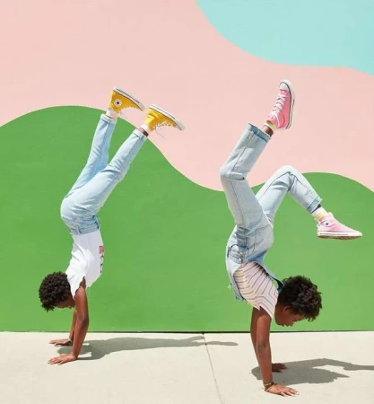 Two children with light skin tones perform handstands outdoors against a colorful wall, wearing blue jeans and yellow and pink sneakers.