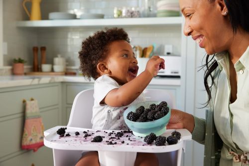 Smiling mother watches her baby in a high chair eating blackberries in a bright kitchen, creating a heartwarming, family moment.